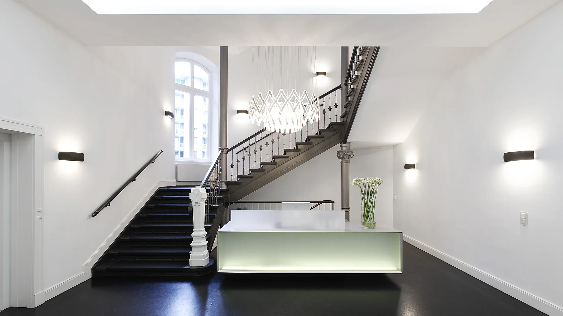 Empfangstresen aus Mineralwerkstoff befindet sich in Eingangshalle eines Altbaus. Ein moderner Kronleuchter erhellt den Raum.