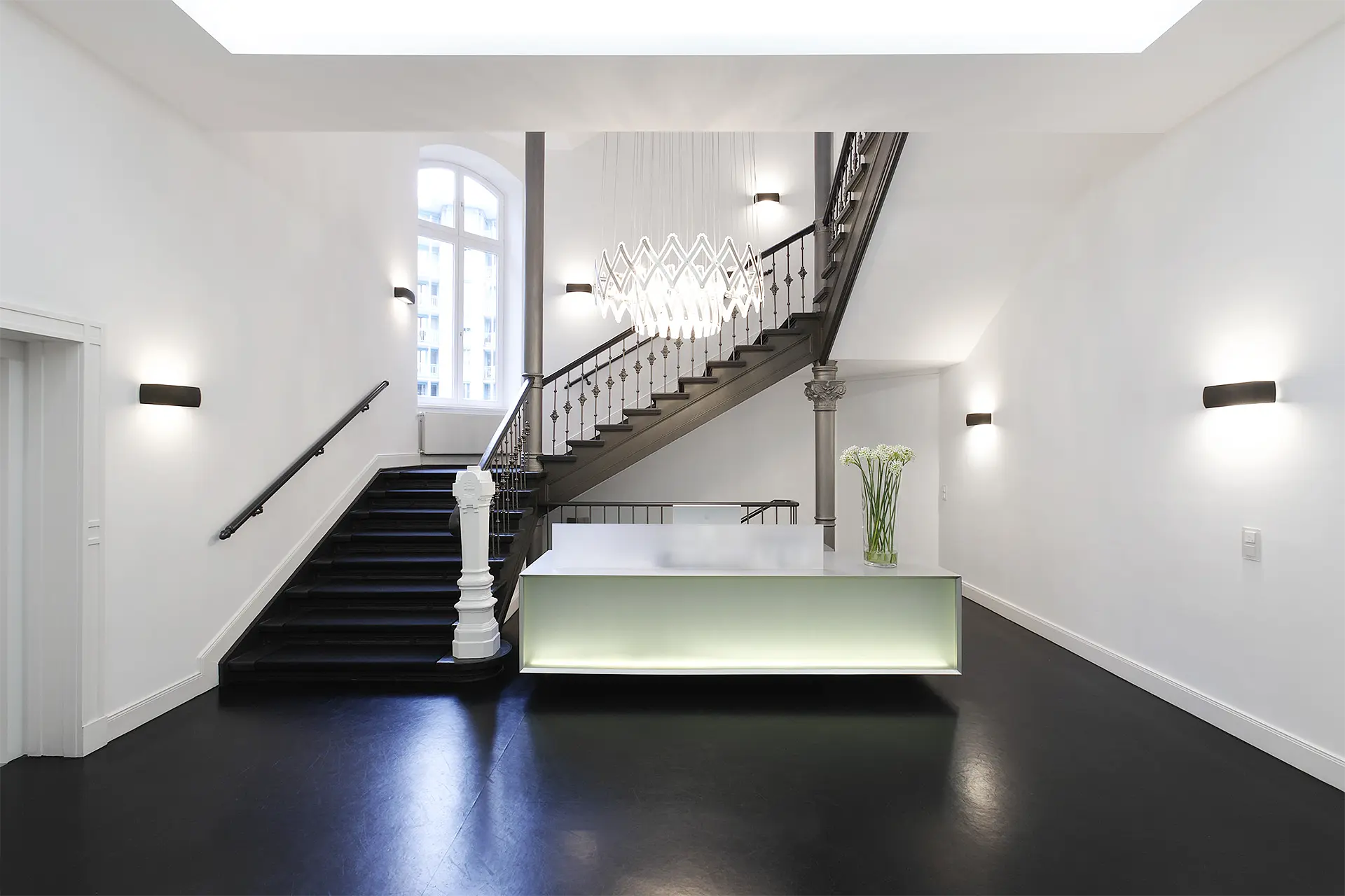 Empfangstresen aus Mineralwerkstoff befindet sich in Eingangshalle eines Altbaus. Ein moderner Kronleuchter erhellt den Raum.