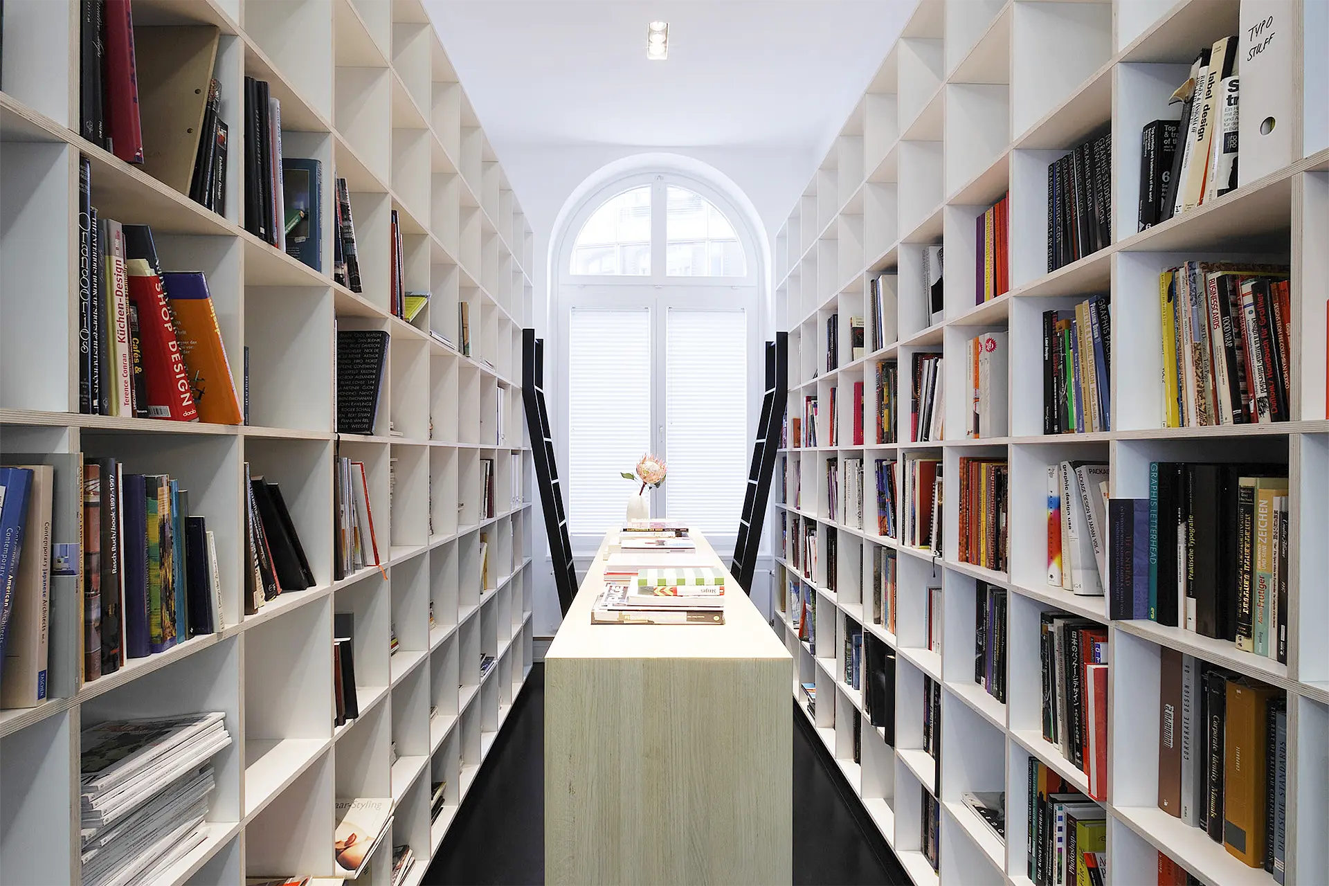 Weiße Regale befinden sich auf beiden Seiten eines Raumes. Die Regalfächer sind mit Büchern befüllt. Zwei schwarze Leiter lehnen an.