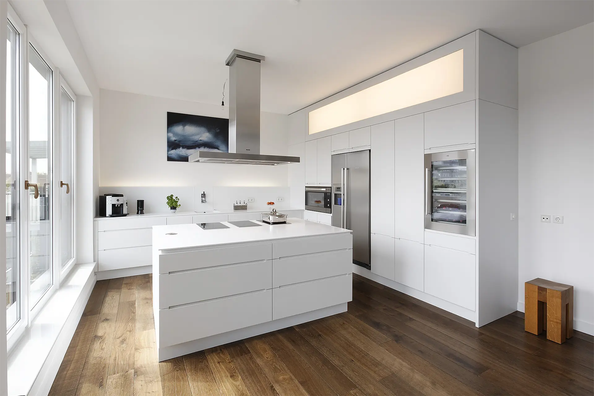 Weiße Küche mit freistehenden Mittelblock in einem Raum mit großen Fenstern.