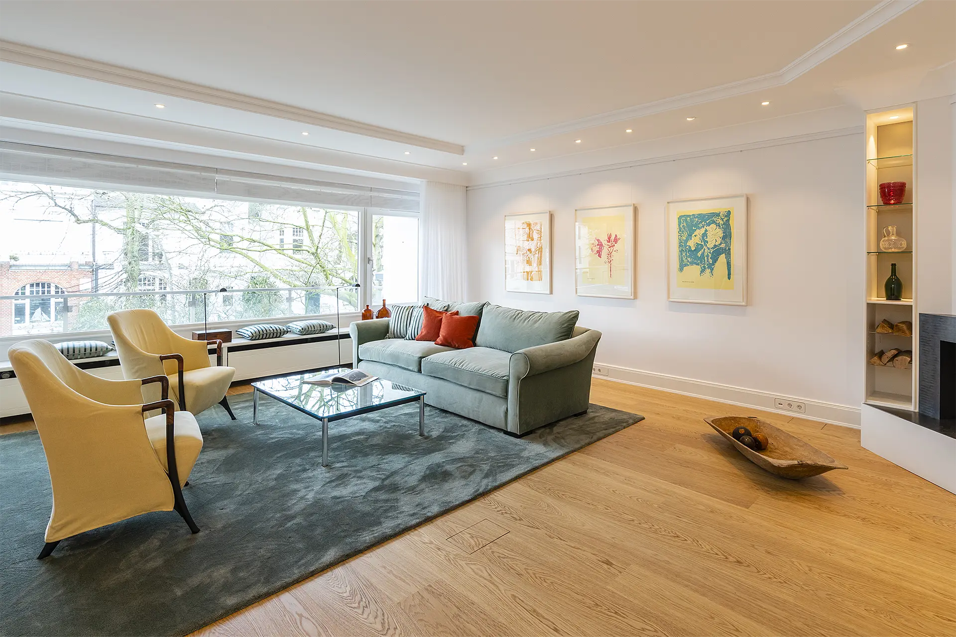 In einem Wohnzimmer mit Fensterfront und mit Kamin stehen ein türkisfarbenes Sofa, zwei gelbe Sessel und ein Glastisch.