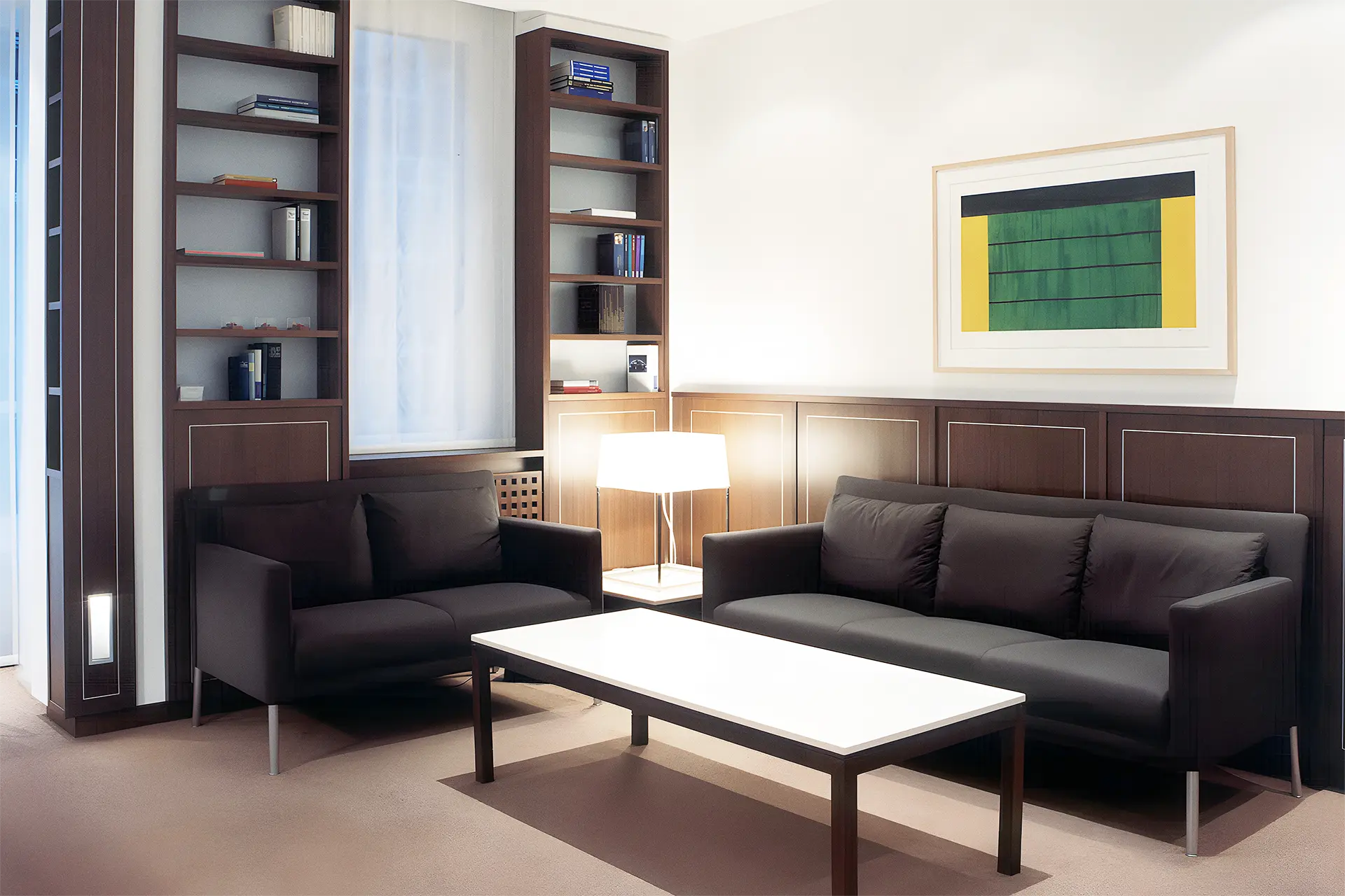 In einen kleinen Konferenzraum befinden sich zwei dunkelbraune Sofaelemente, Tisch, Holzregale, Tischlampe und ein Wandbild.